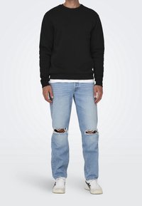 Felpa nera a collo rotondo sopra una camicia bianca, abbinata a jeans strappati blu chiaro e sneaker bianche con accenti neri.