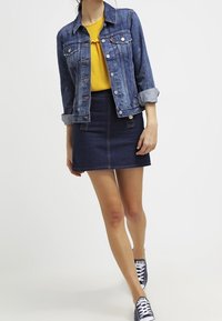 Giacca di denim, camicia gialla con volant, mini gonna di denim scuro e sneaker blu navy. La giacca ha bottoni argentati e maniche arrotolate.