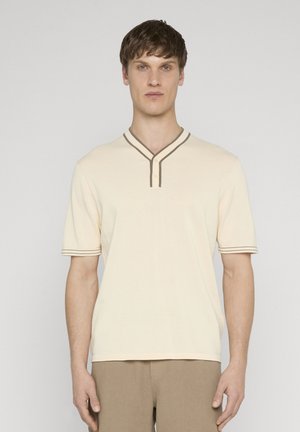 Young man wearing a cream short-sleeve shirt with brown trim on collar and sleeves, paired with light brown pants, standing against plain background.