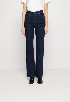 Person iført højtaljede mørkeblå jeans med lige ben, hvid kortærmet top og sorte ankelstøvler med firkantet tå, stående på en hvid gulv.