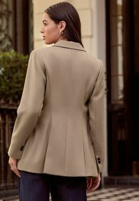 Blazer bej croită, cu un design structurat, guler cu creastă și spate plisat. Detalii includ nasturi negri pe mâneci și material texturat.