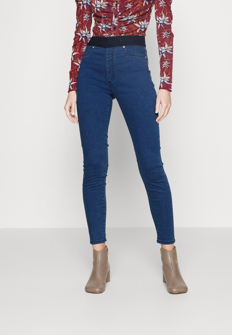 Femme portant un jean skinny bleu taille haute, des bottines marron et une chemise rouge à fleurs à manches longues sur un fond uni.
