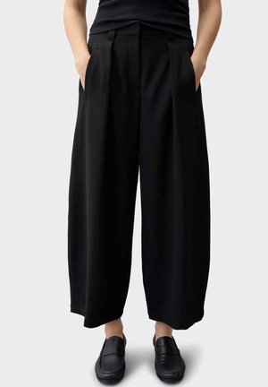 Persona che indossa pantaloni neri a gamba larga con pieghe, mocassini neri e un top nero senza maniche, mani nelle tasche.