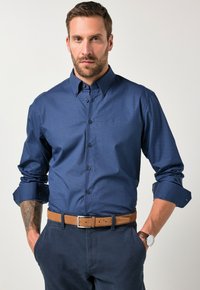 Hombre con barba y un tatuaje en el antebrazo izquierdo, vistiendo una camisa de manga corta azul marino, cinturón marrón, pantalones azul marino y reloj de muñeca, con las manos en los bolsillos.
