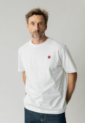 Mann mit kurzen braunen Haaren und Bart, der ein weißes T-Shirt mit einem kleinen roten Kreis auf der Brust trägt, steht vor einem einfarbigen grauen Hintergrund.