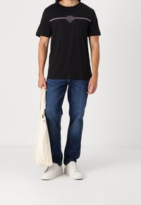 Mand iført sort Tommy Hilfiger T-shirt, blå jeans, hvide sneakers, holder en beige tote bag, står foran en ensfarvet baggrund.