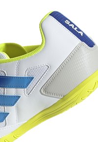Zapatilla deportiva blanca con rayas y acentos azules, suela de neón amarilla, parte superior texturizada y un talón reforzado con la etiqueta "SALA".