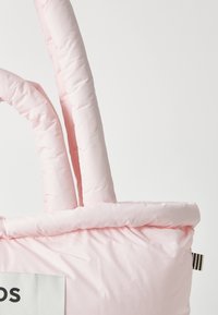 Sac fourre-tout rose rembourré avec deux poignées matelassées, arborant une étiquette en tissu blanc avec du texte noir. Texture douce et design minimaliste.