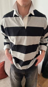 Person wearing a black and gray horizontal striped rugby shirt over a striped collared shirt, standing with hands in jeans pockets indoors.