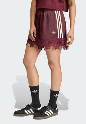 Dolná časť tela osoby nosiacej bordové šortky s čipkovým lemovaním, čierne ponožky Adidas a čierne tenisky Adidas s bielymi pruhmi.