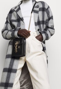 Harmaa ruutukuvioinen villatakki mustilla napilla, käytetty valkoisen poolsikaulan alla. Valkoiset korkeavyötäröiset housut. Musta nahkalaukku kultaisilla yksityiskohdilla.