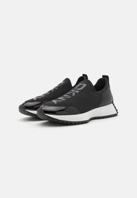Sorte slip-on joggesko med en glatt tekstiloverdel, blanke sorte detaljer, en hvit såle og en teksturert gummisåle.