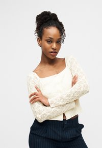 Νεαρή γυναίκα με σγουρά μαλλιά σε ψηλό κότσο, που φοράει λευκό δαντελένιο τοπ με μακριά μανίκια και μπλε παντελόνι με ρίγες, στέκεται με σταυρωμένα τα χέρια.