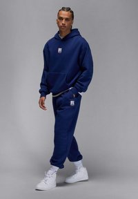 Ung person med fletninger iført en marineblå Jordan-hoodie og matchende joggingbukser, hvide sneakers, stående mod en grå baggrund.