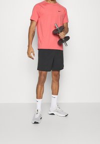 Camiseta deportiva roja de Nike, pantalones cortos negros, calcetines blancos de Nike y zapatillas grises sosteniendo mancuerna. Diseño simple, textura de tela ligera.