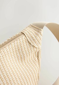 Zbliżenie na beżową, plecioną torbę z zamkiem błyskawicznym i dopasowanym paskiem na jednolitym jasnym tle.