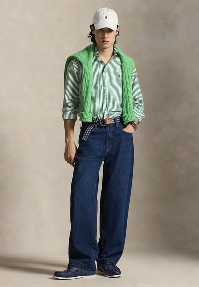 Jeune homme portant une casquette blanche, une chemise rayée verte, un pull vert drapé sur les épaules, un jean large bleu et des chaussures bleues, debout devant un fond neutre.