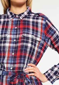 Chemise à carreaux avec un mélange de couleurs rouge, marine et blanche, ouverture boutonnée à l'avant, deux poches poitrine et un détail noué à la taille. Matière en coton.