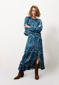 Langes türkisfarbenes Kleid mit weißen abstrakten Mustern, ausgestattet mit einem seitlichen Schlitz, kombiniert mit braunen Cowboystiefeln. Weicher Stoff, lässige Passform und lange Ärmel.