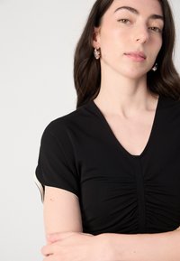 Black top with a V-neck and short sleeves. Features gathered fabric at the front and cream accents on the sleeve edges. Smooth texture.