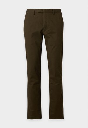 Homme debout portant un t-shirt corail à manches courtes, un pantalon marron foncé et des baskets blanches sur un fond uni clair.