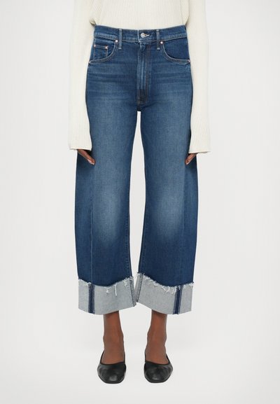 Jeans z wysokim stanem i szerokimi nogawkami w ciemnoniebieskim dżinsie z postrzępionymi szarymi detalami na końcówkach oraz minimalnymi akcentami metalowymi. Gładka tekstura.