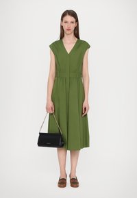 Robe verte, longueur genou, avec un décolleté en V et des manches courtes, cintrée à la taille. Associée à une pochette noire et des chaussures slip-on marron.