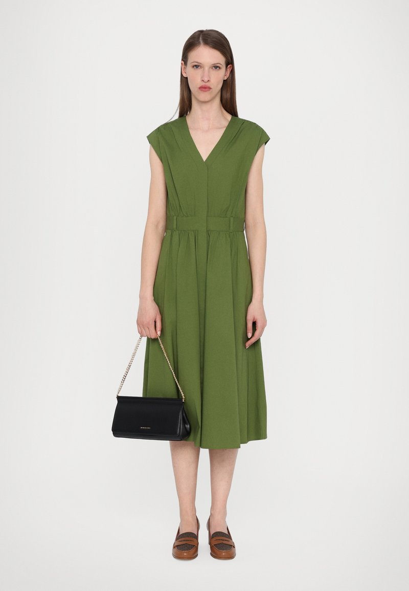 Robe verte, longueur genou, avec un décolleté en V et des manches courtes, cintrée à la taille. Associée à une pochette noire et des chaussures slip-on marron.