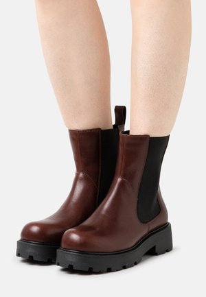 Brune læderankelstøvler med sorte elastiske sidepaneler, en rund tå og en tekstureret sort gummisål med mønster.