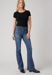 Must mustriga lühikeste varrukatega soonikust top ja sinised laiade lahendustega teksapüksid, millel on keskmise kõrgusega vöökoht ja viie taskuga disain, kantud mustade saapadega.