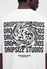 Uomo che indossa una maglietta bianca con una spirale astratta nera sopra il testo ripetuto "DROPSIZE STUDIOS" e la scritta "CREATED IN-HOUSE LIMITED EDITION" sotto.