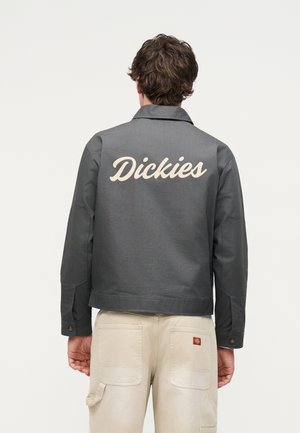 Homme portant une veste Dickies gris foncé avec un logo dans le dos et un pantalon beige clair avec une poche à outils, debout dos à un fond uni.