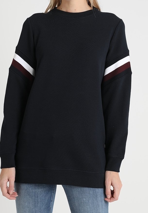 Sudadera negra de manga larga con rayas en blanco y rojo oscuro en la parte superior de los brazos, combinada con jeans azules, vista frontal, sin cabeza visible.