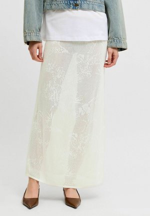 Jupe maxi blanche semi-transparente en maille portée avec un top blanc, une veste en jean et des chaussures marron à pointe.