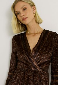 Vestido de estampado de leopardo marrón con un escote en V profundo, mangas largas y cintura fruncida. Presenta acentos dorados en la joyería y un detallado intrincado.