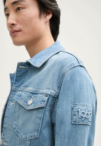 Denim jakna svijetloplave boje s dugmadi sprijeda, dva džepa na prsima i teksturiranim aplikacijom s "1962" na rukavu.