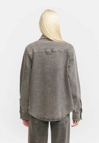 Persoon met lang blond haar, gekleed in een losse, vervaagde grijze denimjas en bijpassende broek, met de rug naar de camera tegen een effen achtergrond.