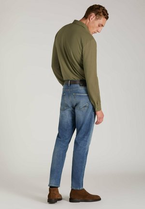 Olivgrünes Langarmhemd mit einem knöpfbaren Kragen, kombiniert mit hellblauen Jeans. Braune Wildleder-Stiefeletten.