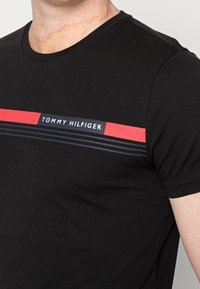 Camiseta de algodón negra con cuello redondo y mangas cortas. En la parte frontal presenta un diseño a rayas en rojo y azul marino con el logo "TOMMY HILFIGER".