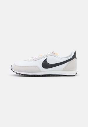 Hvid og lysegrå Nike løbesko med sort swoosh-logo og tekstureret sort sål, vist fra siden på hvid baggrund.