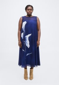 Γυναίκα που φοράει μια αμάνικη μεσάτη μπλε φορέμα με μεγάλο λευκό ανθισμένο σχέδιο, συνδυασμένη με ταμπά δερμάτινα μποτάκια αστραγάλου.