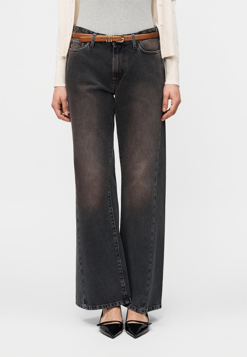 Γυναίκα που φοράει ξεθωριασμένο μαύρο τζιν με φαρδιά μπατζάκια και καφέ ζώνη, λευκό πουλόβερ, κρεμ ζακέτα και μαύρες γόβες με μυτερή μύτη, στέκεται μπροστά σε απλό φόντο.