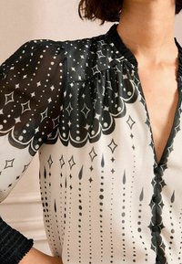 Svart och vit blus med V-ringning, som har genomskinliga puffärmar, intrikata mönster av stjärnor och prickar samt detaljerad plissering nära halslinningen.