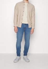 Camisa beige claro abotonada sobre una camiseta crema, combinada con jeans ajustados azules y zapatillas grises. La camisa tiene un bolsillo en el pecho.