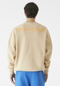Beige sweatshirt med en avslappet passform, med oversized oransje tekst "9N1M SENSE." på baksiden. Ribbestrikkede mansjetter og kant. Kombinert med blå bukser.