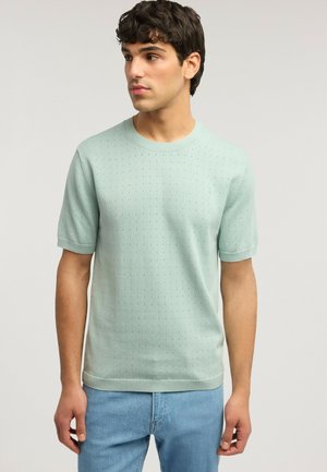 Jeune homme portant une chemise en maille à manches courtes vert pâle avec de petites perforations et un jean bleu clair, regardant légèrement vers la gauche.