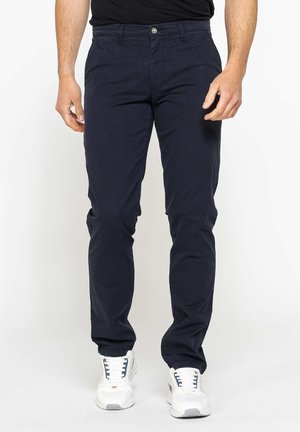Uomo con pantaloni blu scuro e scarpe da ginnastica bianche in piedi davanti a uno sfondo bianco.