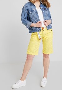 Veste en jean aux manches retroussées, t-shirt blanc et short jaune jusqu'aux genoux, associés à des baskets blanches. Le short jaune est ceinturé.