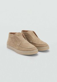 Beige mockasinsanklar med en slip-on design, mjuk textur, rund tå och en låg gummisula. Har en bred, distinkt klaff fram.