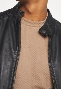 Giacca di pelle nera con chiusura a zip e collo a scatto, abbinata a un top beige strutturato con motivo a spina di pesce.